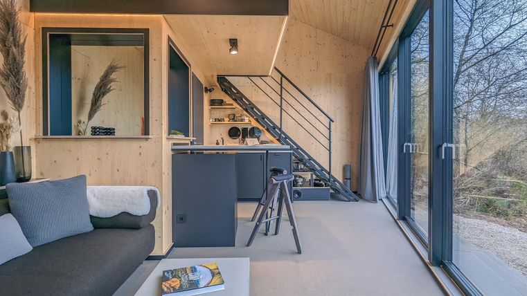 Ein modernes, minimalistisches Wohnzimmer mit großen Fenstern und einer Treppe. Die Einrichtung besteht aus Holz und schlichten Möbeln.