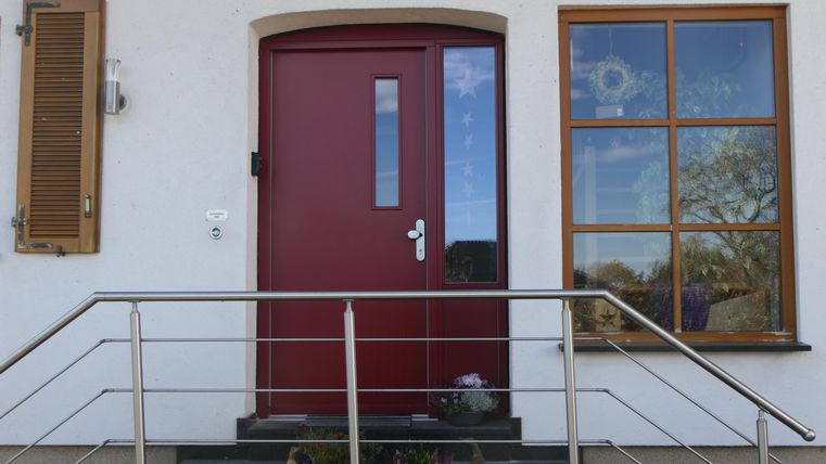 Eine rote Haustür mit Glaselementen und ein Fenster nebenher. Die Eingangsbereich ist mit einer modernen Treppe und Blumen dekoriert.