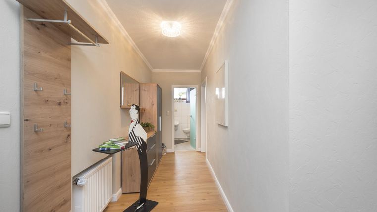 Ein heller Flur mit Holzregalen und modernem Mobiliar. Am Ende des Flurs ist eine Tür zu sehen, die zu einem weiteren Raum führt.