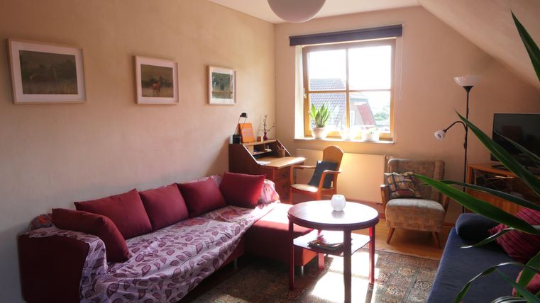 Ein gemütliches Wohnzimmer mit einem roten Sofa und einem runden Tisch. Das Zimmer hat viel Tageslicht und Pflanzen am Fenster.