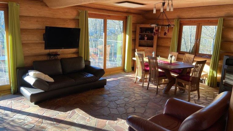 Ein gemütliches Wohnzimmer mit Holzverkleidung, einem großen Fenster und Blick auf die Natur. Es gibt ein Sofa, einen Essbereich mit Stühlen und einen Fernseher.