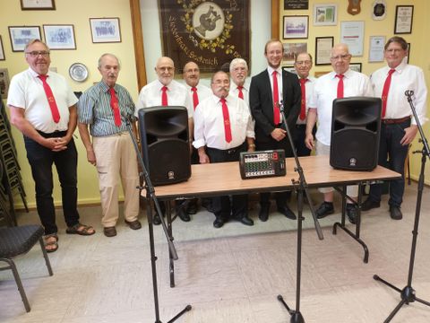 Eine große Blaskapelle steht im Freien und posiert für ein Gruppenfoto. Die Musiker tragen farbige Uniformen und halten ihre Instrumente.