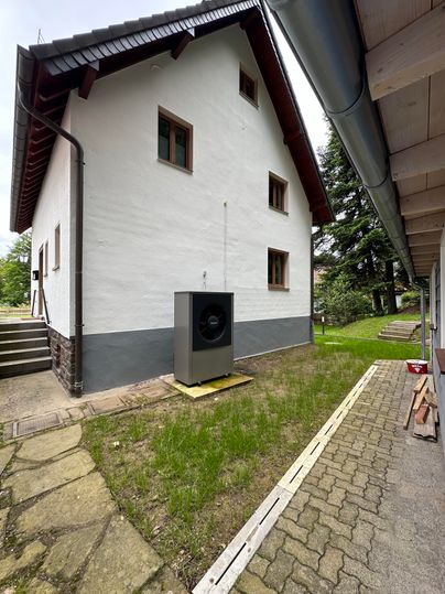 Ein mehrstöckiges Haus mit weißer Fassade und braunen Fenstern. Im Vordergrund befindet sich eine Wärmepumpe auf einem kleinen Podest.