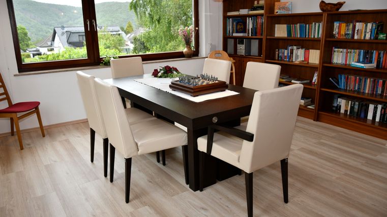 Ein gemütlicher Essbereich mit einem Holzstuhl und einem großen Tisch, umgeben von beigen Stühlen. Im Hintergrund sieht man Regale mit Büchern und Fenster mit Blick auf die Landschaft.