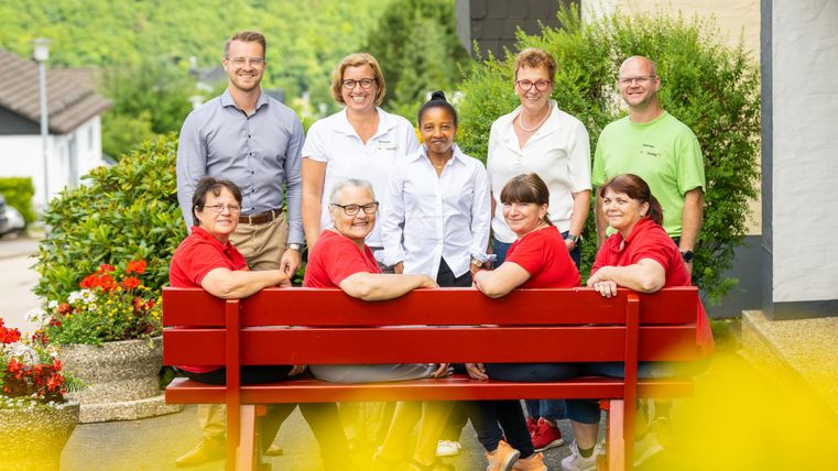 Eine Gruppe von zehn Personen steht und sitzt auf einer roten Bank vor einem Gebäude. Die Umgebung ist grün mit Blumen und einem klaren Himmel.