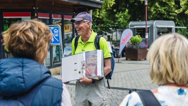 Ein älterer Mann mit einer Sonnenbrille und einem grünen Hemd hält einen Ordner in der Hand. Er spricht mit zwei Frauen in der Stadt.