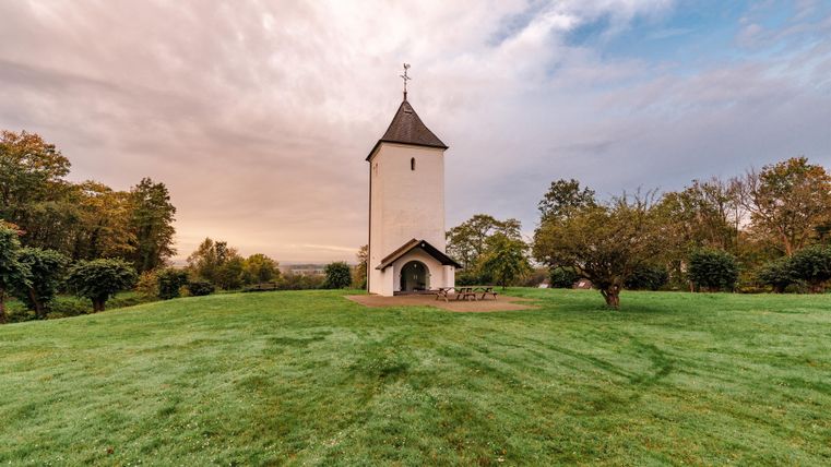 Eine kleine Kirche steht auf einer grünen Wiese. Im Hintergrund sind Bäume und ein sanfter Himmel zu sehen.