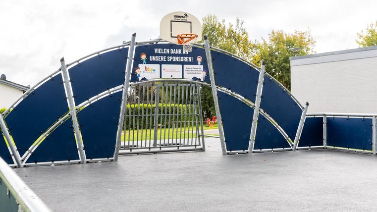 Ein Basketballplatz mit einem halbkreisförmigen Korb und einer stabilen Umzäunung. Im Hintergrund ist ein grünes Gelände und ein Gebäude zu sehen.