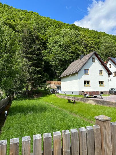 Ein gemütliches Haus steht am Rande eines Waldes. Der Garten ist grün und gut gepflegt, umgeben von Bäumen und einem blauen Himmel.