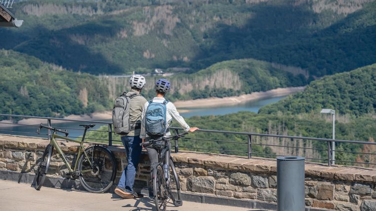 Zwei Radfahrer stehen an einem Aussichtspunkt und betrachten die Landschaft. Im Hintergrund sind sanfte Hügel und ein Gewässer zu sehen.