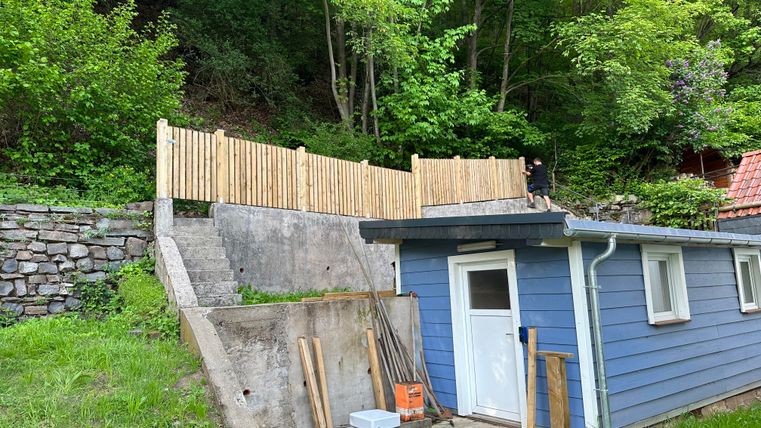 Ein kleines blaues Haus steht am Hang zwischen Bäumen. Im Hintergrund ist ein hölzernes Geländer und eine gepflasterte Fläche zu sehen.