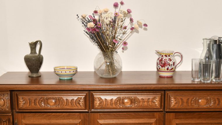 Eine Holzkommode mit dekorativen Teilen. Auf der Oberfläche steht eine Blumenvase mit Trockenblumen, sowie verschiedene Töpfe und Gläser.