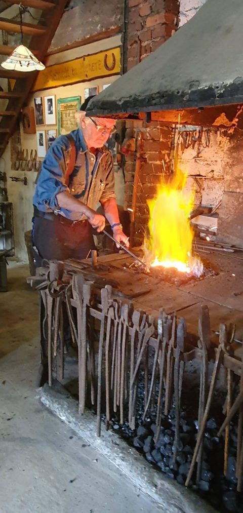 Ein Schmied arbeitet an einem Amboss, während Flammen aus dem Feuerofen lodern. Im Hintergrund sind Werkzeuge und Ausrüstungen sichtbar.