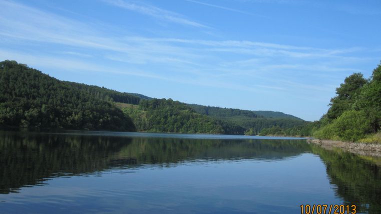 Ein ruhiger See umgeben von grünen Hügeln und klarem Himmel. Das Wasser spiegelt die Landschaft wider.