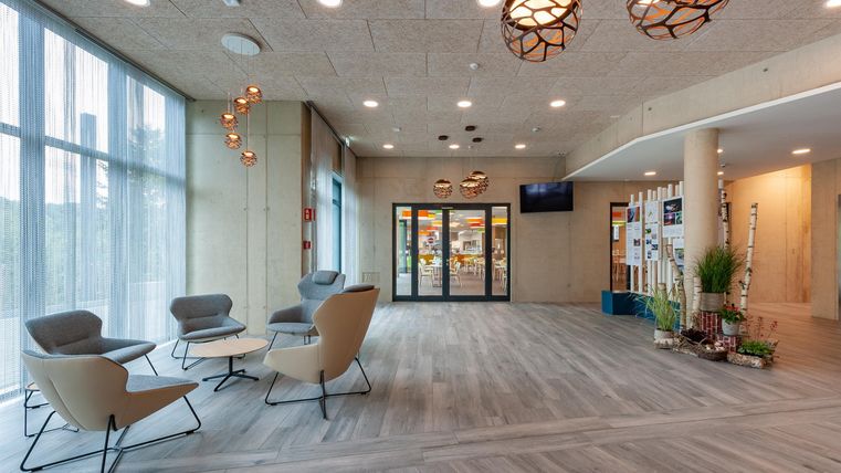 Ein moderner Empfangsbereich mit großen Fenstern und hellem Holzfußboden. Gemütliche Sitzgelegenheiten und stilvolle Lampen sorgen für eine einladende Atmosphäre.