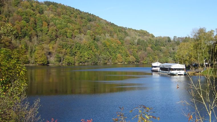 Ein ruhiger See umgeben von buntem Herbstlaub. Zwei Boote liegen friedlich am Ufer.