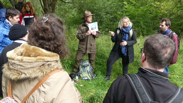 Eine Gruppe von Menschen steht im Freien und hört einer Präsentation zu. Im Vordergrund zeigt eine Person ein Bild, während andere aufmerksam zuhören.