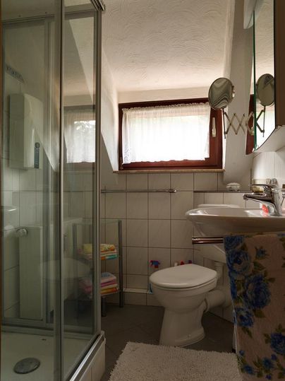 Ein Badezimmer mit Dusche, Waschbecken und Toilette. Es gibt helle Fliesen und ein Fenster, das Tageslicht hereinkommen lässt.