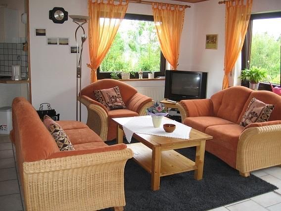 Ein gemütliches Wohnzimmer mit zwei orangenen Sofas und einem Holztisch. Große Fenster lassen viel Licht herein und bieten Blick auf die grüne Landschaft draußen.