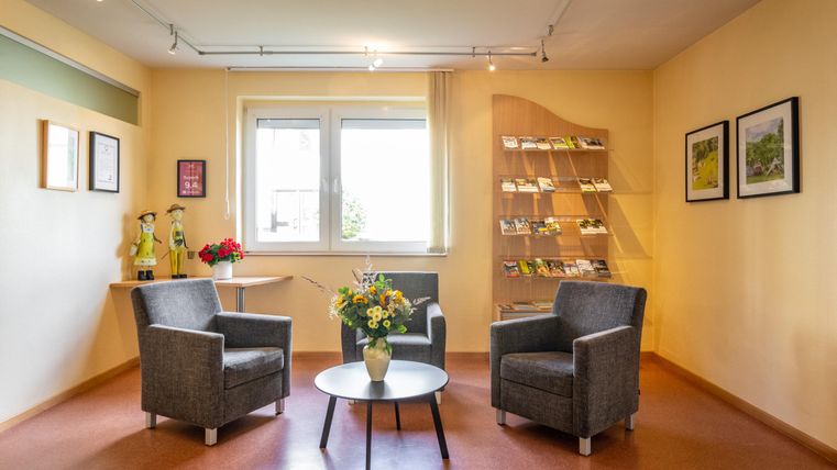 Ein gemütlicher Raum mit zwei grauen Sesseln und einem runden Tisch. An der Wand sind Broschüren und einige Bilder zu sehen.