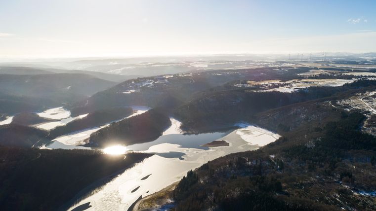 Eine malerische Landschaft mit sanften Hügeln und einem glitzernden Gewässer. Der Himmel ist klar und die Sonne scheint hell.