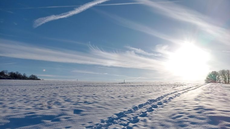 Eine verschneite Landschaft unter einem klaren blauen Himmel. Die Sonnenstrahlen scheinen hell und warme Fußspuren führen durch den Schnee.