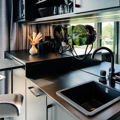 Eine moderne Küche mit dunklen Oberflächen und einem eleganten Waschbecken. Großflächige Fenster sorgen für viel natürliches Licht und Ausblick ins Grüne.