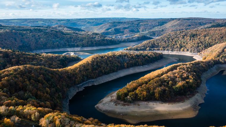Eine malerische Landschaft mit sanften Hügeln und einem geschlängelten Fluss. Die Bäume leuchten in herbstlichen Farben unter einem klaren Himmel.